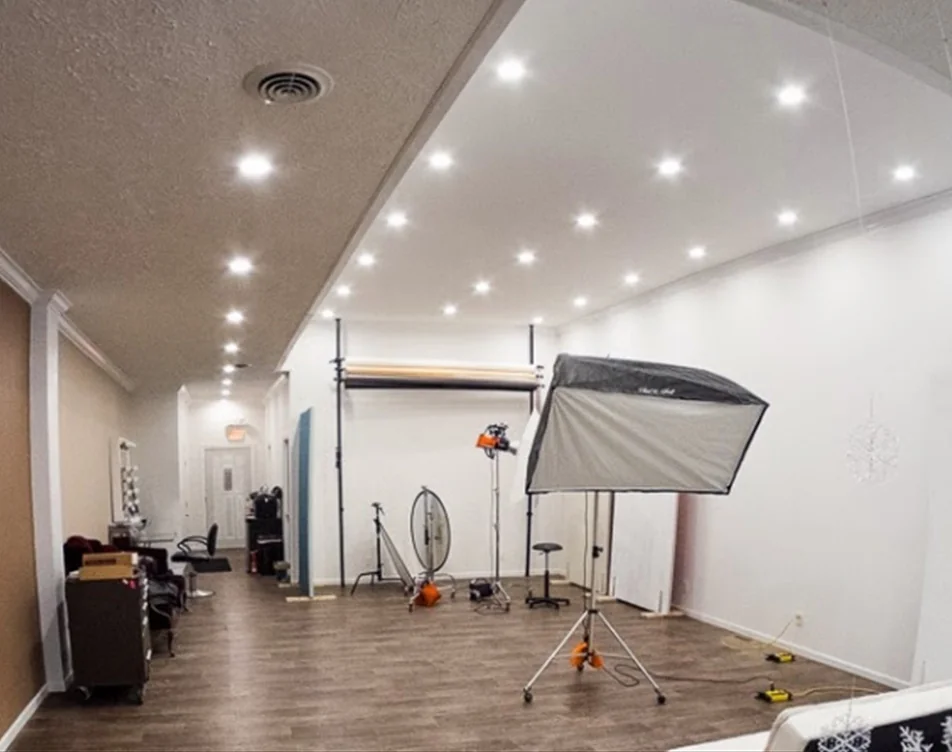 The main shooting space at Studio M: a softbox on a stand, rolls of backdrop paper on a wall bar, and a bank of recessed ceiling lights over a wood floor